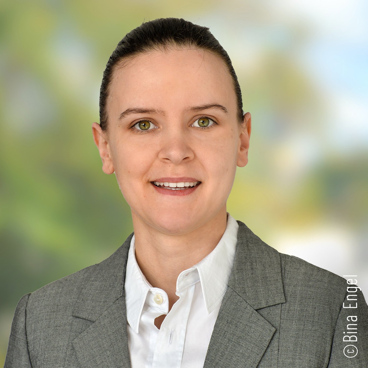 Eine Person mit heller Haut, rötlichem Haar und Brille lächelt, trägt ein weißes Hemd und einen dunklen Blazer vor einem unscharfen grünen Hintergrund im Freien.