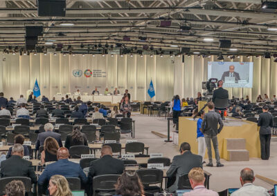 Frontalblick auf eine Panel-Bühne auf dem Gelände der Konferenz. Ein Speaker ist auf einer Leinwand zu sehen, zahlreiche Gäste mit Namensschildern hören zu.
