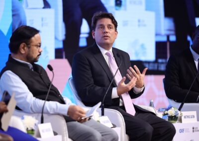 Vier Männer in Geschäftskleidung sitzen auf einer Podiumsdiskussionsbühne, jeder mit Mikrofonen und Notizen, während ein Mann in der Mitte spricht und mit seinen Händen gestikuliert. Hinter ihnen befindet sich ein großer blau-weißer Veranstaltungshintergrund.