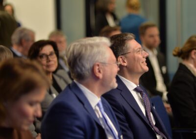 Menschen in Geschäftskleidung sitzen bei einer Indoor-Veranstaltung eng beieinander, wirken beschäftigt und lächeln. Der Fokus liegt auf zwei Männern in blauen Anzügen im Vordergrund, umgeben von anderen Teilnehmern im unscharfen Hintergrund.