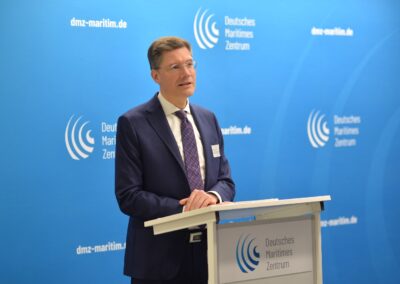 Ein Mann in Anzug und Krawatte steht an einem Podium und spricht vor einem blauen Hintergrund mit dem Logo und Text des Deutschen Maritimen Zentrums. Es scheint sich um eine formelle Veranstaltung oder Pressekonferenz zu handeln.