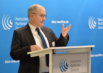 Ein Mann in Anzug und Krawatte spricht an einem Podium mit dem Deutschen Maritimen Zentrum und maritimen Logos auf blauem Hintergrund.