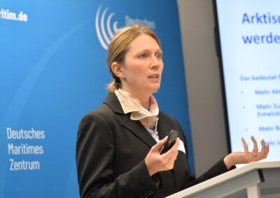 Eine Frau in einem schwarzen Anzug spricht an einem Podium mit einem Mikrofon in der Hand vor einem blauen Hintergrund mit der Aufschrift Deutsches Maritimes Zentrum. Neben ihr ist ein Bildschirm mit deutschem Text zu sehen.