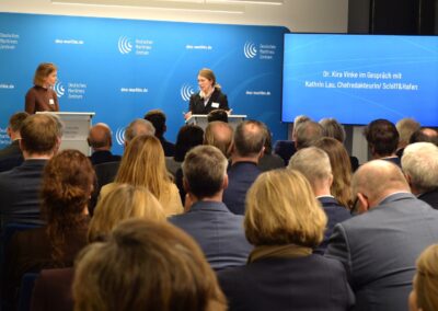 Eine Frau spricht von einem Podium aus zu einem Publikum, während eine andere Frau daneben steht und einen Ordner hält. Hinter ihnen sind ein blauer Hintergrund und ein Bildschirm zu sehen, auf dem die Namen Dr. Kira Vinke und Kathrin Lau stehen.