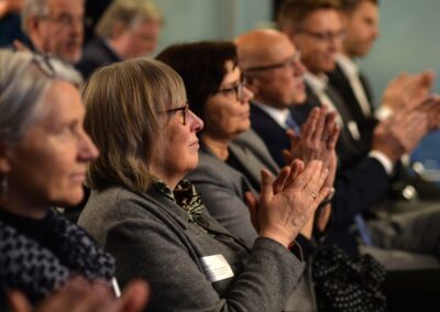 Eine Gruppe Erwachsener sitzt in Reihen und beobachtet aufmerksam und applaudiert etwas aus dem Off. Die meisten sind in Geschäftskleidung gekleidet und wirken konzentriert und engagiert in einer formellen Umgebung.