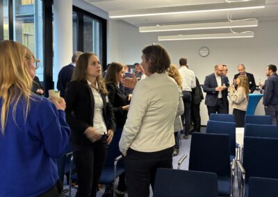 People are mingling and conversing in a modern conference room with rows of empty chairs, large windows, and a clock on the wall. The atmosphere appears casual and social, suggesting a networking or business event.