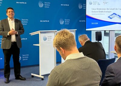 A man in a suit speaks at a podium in front of a blue backdrop. Several people are seated facing him. A screen displays a presentation on the future of onshore wind turbine transport.