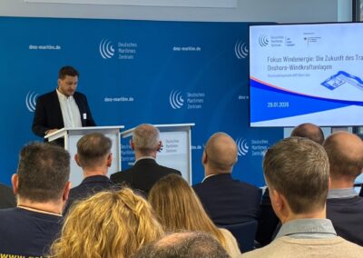 A man stands at a podium presenting to an audience in a conference room. Behind him is a blue backdrop with Deutsches Maritimes Zentrum logos. A large screen displays a presentation on the future of onshore wind turbine transport.