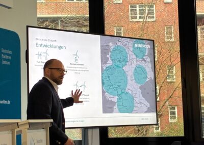 A man in a suit presents a slide about wind energy developments, featuring maps and diagrams, in a modern conference room with large windows showing brick buildings outside.