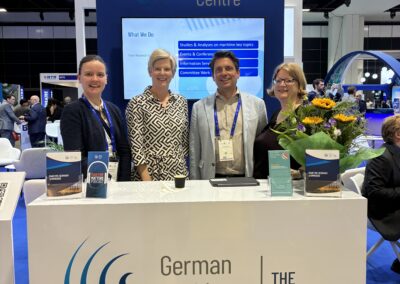 Vier Personen stehen lächelnd hinter einem Stand des German Maritime Centre auf einer Konferenz. Eine Person hält einen Blumenstrauß in der Hand. Der Stand trägt ein blau-weißes Schild mit dem Namen der Organisation und dem Slogan "Der Zukunft auf der Spur".