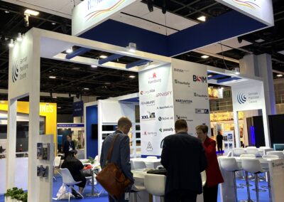 Drei Personen stehen und unterhalten sich an einem Stand mit der Aufschrift "Maritime Germany" auf einer Messe. Der Stand ist mit weißen Möbeln, Firmenlogos an den Wänden und einem blauen Teppichboden ausgestattet. Im Hintergrund sind andere Stände zu sehen.