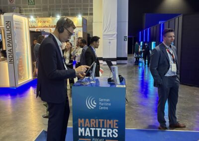 Ein Mann mit Kopfhörern steht an einem Podcast-Stand mit der Aufschrift German Maritime Centre Maritime Matters auf einer Messe, während andere in der Nähe herumlaufen und sich unterhalten. Im Hintergrund sind der indische Pavillon und andere Messestände zu sehen.