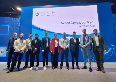 Zehn Personen stehen in einer Reihe auf einer Bühne vor einem großen Bildschirm mit der Aufschrift Maritime Germany awaits you at Booth B16! und dem Logo des German Maritime Centre. Sie sind in Business-Kleidung gekleidet und lächeln in die Kamera.