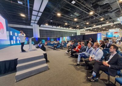 Ein Redner spricht auf der Bühne zu einem aufmerksamen Publikum auf einer Konferenz, wobei ein projiziertes Tortendiagramm auf der Leinwand zu sehen ist. Der Raum ist geräumig, modern beleuchtet und verfügt über einen großen Sitzbereich.