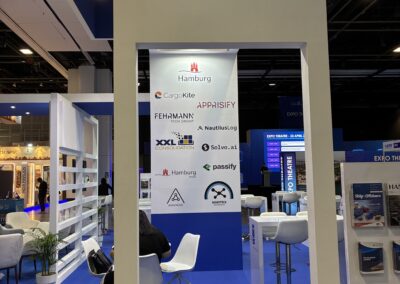 Ein Display am Stand des German Maritime Centre zeigt Logos verschiedener Unternehmen. Der Bereich ist mit blauem Teppichboden, weißen Stühlen, Tischen und Grünpflanzen an der Basis des Standes ausgestattet. Im Hintergrund sitzen und gehen Menschen.