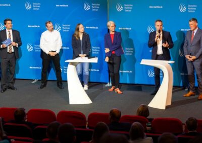 Sechs Personen stehen auf einer Bühne mit blauem Hintergrund und dem Logo "Deutsches Maritimes Zentrum" auf dem DMZ-Side-Event zur 14. Nationalen Maritimen Konferenz. Zwei Redner sprechen zum Publikum, andere hören zu, im Vordergrund sitzen die Teilnehmer.