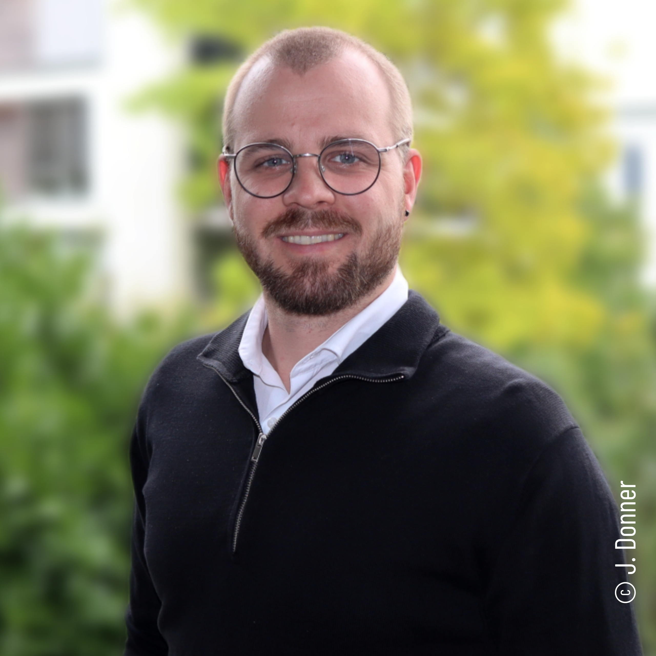 Jan Sebastian Donner Ein lächelnder Mann mit Brille und Bart, der einen schwarzen Pullover mit Reißverschluss über einem weißen Hemd trägt, steht im Freien mit Grünpflanzen und verschwommenen Gebäuden im Hintergrund. © J. Donner.