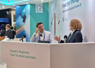 Zwei Männer sitzen an einem Stand mit der Aufschrift Lloyd's Register. Ihr vertrauenswürdiger Berater. auf einer Fachmesse und unterhalten sich. Hinter ihnen befindet sich ein Display mit Bildern und Texten zu Berichten über alternative Kraftstoffe von LR.