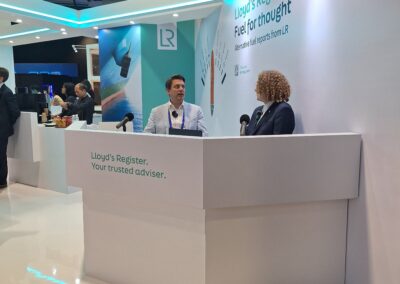 Zwei Personen sitzen an einem modernen weißen Stand, auf dem Lloyd's Register. Your trusted adviser. steht auf der Vorderseite. Hinter ihnen befindet sich ein großes Banner von Lloyd's Register Fuel for thought. Die Umgebung scheint eine Messe oder Konferenz zu sein.