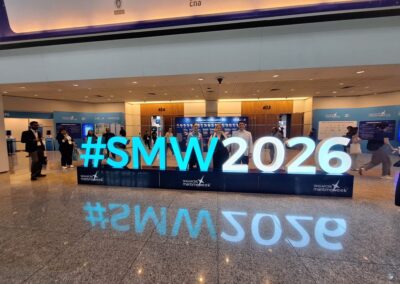 Großes beleuchtetes Schild mit der Aufschrift #SMW2026 an einem Veranstaltungsort in einer Halle mit dahinter stehenden Menschen und Informationsständen im Hintergrund. Der Boden ist reflektierend und die Halle ist hell erleuchtet.