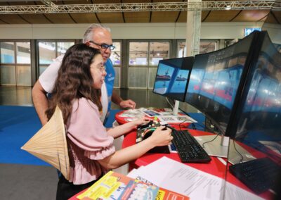 Eine junge Frau und ein älterer Mann bedienen Joysticks und eine Tastatur, während sie bei einer Indoor-Veranstaltung auf drei großen, gebogenen Monitoren ein Simulationsspiel spielen. Auf dem Tisch neben ihnen liegen Broschüren und Unterlagen.