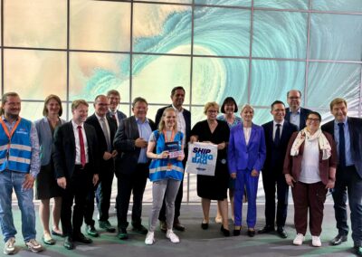 Eine Gruppe von Menschen, von denen einige Business-Kleidung tragen und andere blaue Westen, steht lächelnd in einem Raum vor einem großen digitalen Bildschirm, auf dem eine Meereswelle zu sehen ist. Zwei Personen ganz vorne halten ein Schild mit der Aufschrift „SHIP FLAG“ hoch.