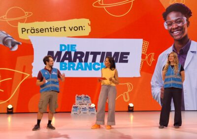 Drei Personen stehen auf einer hell beleuchteten Bühne vor einem orangefarbenen Hintergrund. Zwei von ihnen tragen blaue Westen, und eine hält ein Klemmbrett in der Hand. Hinter ihnen ist der Schriftzug „Präsentiert von: Die Maritime Branche“ zu sehen, begleitet von Grafiken zum Thema Wissenschaft.