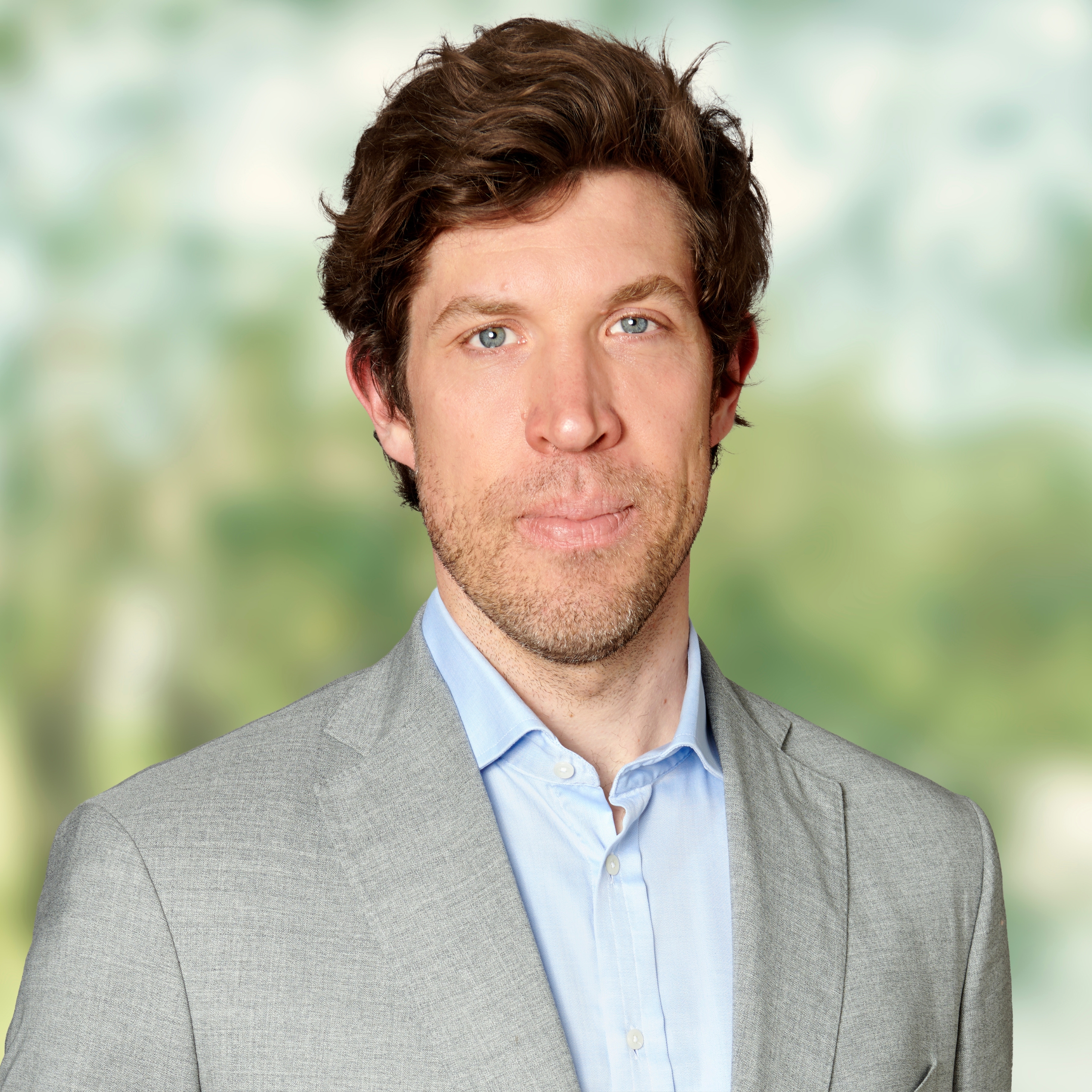 Ein Mann mit welligem braunem Haar und leichtem Bartstoppeln trägt ein hellgraues Sakko über einem hellblauen Hemd und steht vor einem verschwommenen grünen Hintergrund im Freien.
