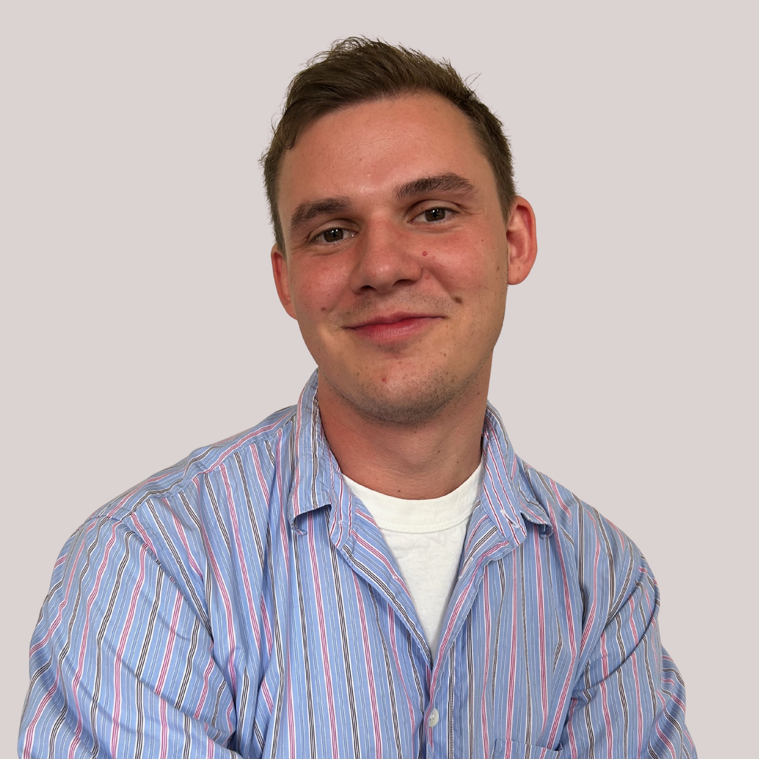 Behler_Timm_Trendscout2 Ein junger Mann mit kurzen braunen Haaren lächelt sanft in die Kamera. Er trägt ein blau-rosa gestreiftes Hemd mit Knöpfen über einem weißen T-Shirt vor einem neutralen hellgrauen Hintergrund.