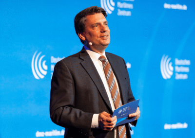 A man in a suit and striped tie speaks on stage, holding a blue folder, with a microphone on his cheek and a blue background displaying logos and circular graphics.
