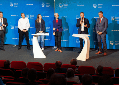 Six people onstage in business attire participate in a panel discussion with microphones, standing at podiums in front of a blue backdrop as an audience watches from red seats.
