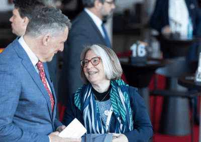 A man in a suit and a woman with glasses and a scarf are smiling and talking at a formal event with other people and tables in the background.