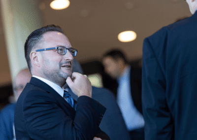 A man in a suit and glasses stands indoors, thoughtfully touching his chin, while talking to another person in a suit. Other people are blurred in the background.