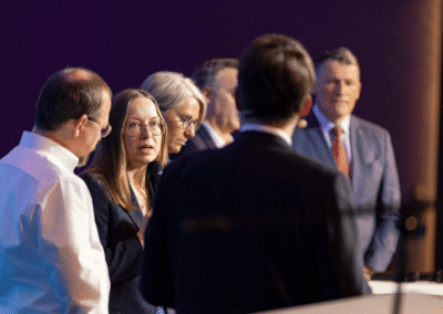 Six adults stand together having a discussion on stage. One woman in the center speaks into a microphone while the others listen attentively. They are dressed in business attire, with a dark background behind them.