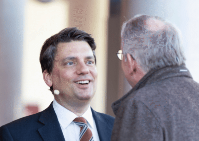 A man in a suit and tie with a headset microphone smiles while speaking to another man, who is seen from behind and wearing glasses and a brown jacket.