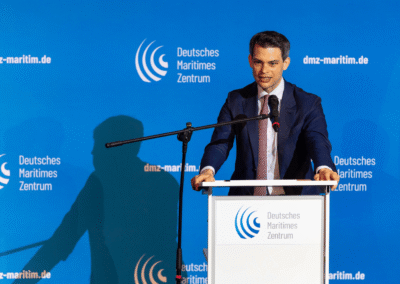 A man in a suit speaks at a podium with the Deutsches Maritimes Zentrum logo, against a blue background with the same logo and website dmz-maritim.de visible.