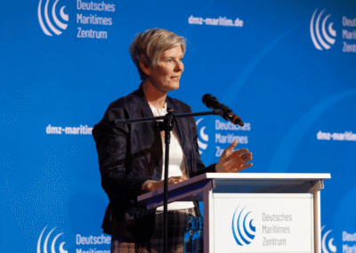 A person stands at a podium speaking into a microphone, with a blue background displaying the Deutsches Maritimes Zentrum logo and text.