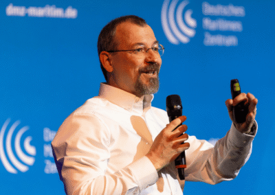 A man in glasses and a white shirt is speaking on stage, holding a microphone and a remote. The blue background displays the logo and text of the Deutsches Maritimes Zentrum.