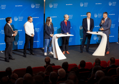 Six people stand on stage with microphones, participating in a panel discussion in front of a blue backdrop with “Deutsches Maritime Zentrum” logos, while an audience watches from red seats.
