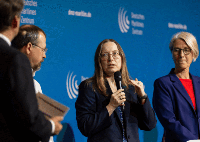 Four people in business attire stand in front of a blue backdrop. A woman in the center speaks into a microphone while others listen. The backdrop has white logos and German text.