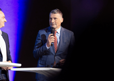 Two men in suits stand on stage. One man speaks into a microphone while the other smiles and looks at him. They are standing behind white tables with a blue and black background.