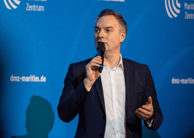 A man in a dark suit holds a microphone and speaks in front of a blue backdrop with white text and logos, including dmz-maritim.de and Deutsches Maritimes Zentrum.