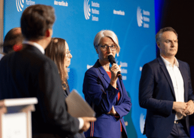 Five people stand on stage at a forum. A woman with short gray hair speaks into a microphone while others listen. The background is blue with white text and logos.