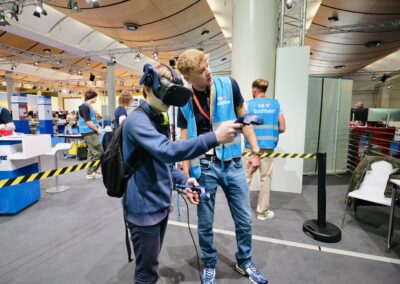 Eine Person, die ein VR-Headset trägt und Controller in den Händen hält, probiert ein Virtual-Reality-Erlebnis aus, während eine andere Person in einer blauen Weste sie durch einen Veranstaltungsraum mit Ständen und heller Beleuchtung führt.