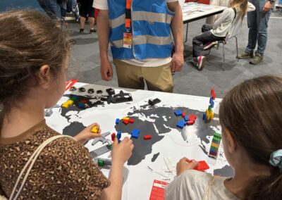 Zwei Kinder bauen mit bunten LEGO-Steinen auf einem Weltkartentisch, während ein Erwachsener in einer blauen Weste daneben steht. Im Hintergrund sind weitere Personen zu sehen, die ähnlichen Aktivitäten nachgehen.