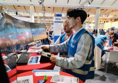 Zwei junge Männer in blauen Freiwilligenwesten bedienen Bedienfelder und blicken auf mehrere gebogene Computermonitore bei einer Veranstaltung in einer Halle, im Hintergrund sind weitere Personen und Stände zu sehen.