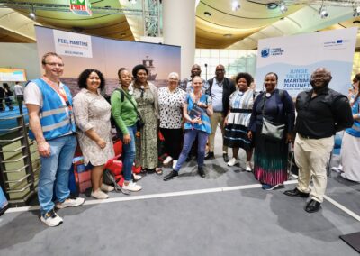 Eine bunt gemischte Gruppe von Menschen steht bei einer Veranstaltung in einem Raum beieinander und lächelt in die Kamera. Einige tragen blaue Westen. Im Hintergrund sind Ausstellungsbanner und Informationsstände zum Thema maritime Berufe zu sehen.