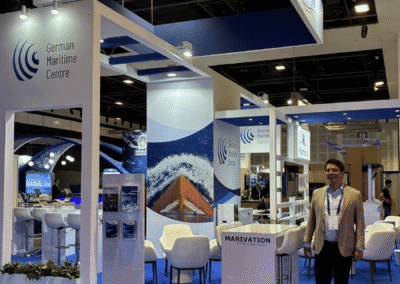 Messestand für „Maritime Germany“ mit blauem Teppichboden, weißen Stühlen und Bannern. Ein Mann in einer beigen Jacke steht am Stand. Die Ausstellung zeigt Bilder zum Thema Seefahrt sowie Logos des Deutschen Maritimen Zentrums.