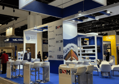 Messestand mit „Maritime Germany“-Schildern, blauem Bodenbelag, weißen Stühlen und Tischen sowie Ausstellungstafeln mit den Logos verschiedener Unternehmen. Menschen laufen durch die Messehalle.