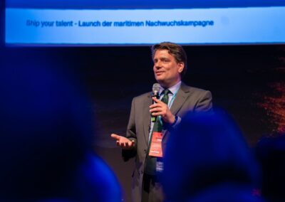 Ein Mann im Anzug spricht bei einer Veranstaltung mit einem Mikrofon auf der Bühne. Er lächelt, hält eine Notizkarte in der Hand und trägt ein rotes Schlüsselband. Im Vordergrund sind blaues Licht und verschwommene Zuschauer zu sehen.